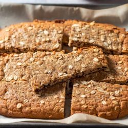 Golden Crunchies Recipe – Buttery Oats, Coconut & Golden Syrup - Saffarazzi Oat-topped baked bars cut into squares on a parchment-lined baking sheet.