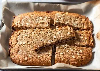 Golden Crunchies Recipe – Buttery Oats, Coconut & Golden Syrup - Saffarazzi Oat-topped baked bars cut into squares on a parchment-lined baking sheet.