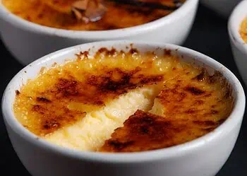 Crème brûlée in white ramekins with a caramelized sugar crust; a spoon-sized crack reveals creamy custard inside.