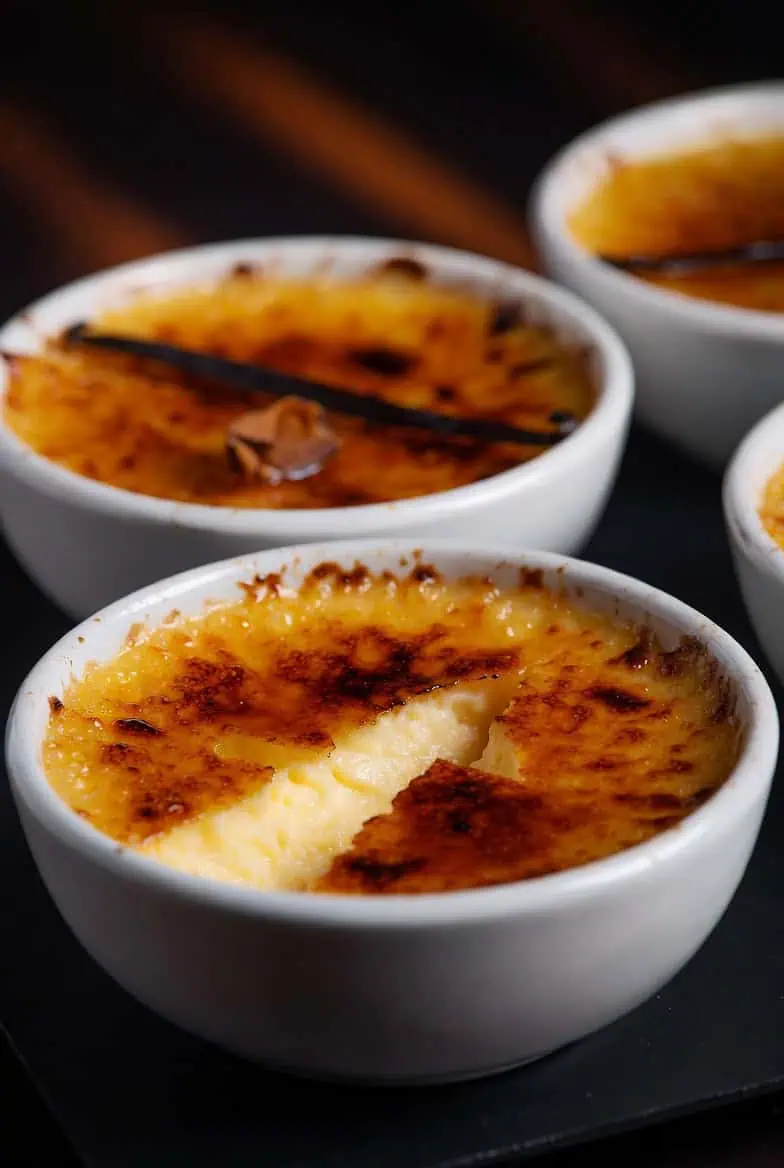 Crème brûlée in white ramekins with a caramelized sugar crust; a spoon-sized crack reveals creamy custard inside. - Saffarazzi Crème brûlée in white ramekins with a caramelized sugar crust; a spoon-sized crack reveals creamy custard inside.