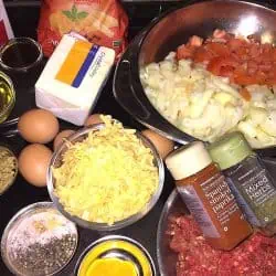 Ingredients Macaronie Mince - Saffarazzi A kitchen prep station with eggs, shredded cheddar, diced onion, chopped tomatoes, blocks of cheese, oil, and assorted spices ready for cooking.