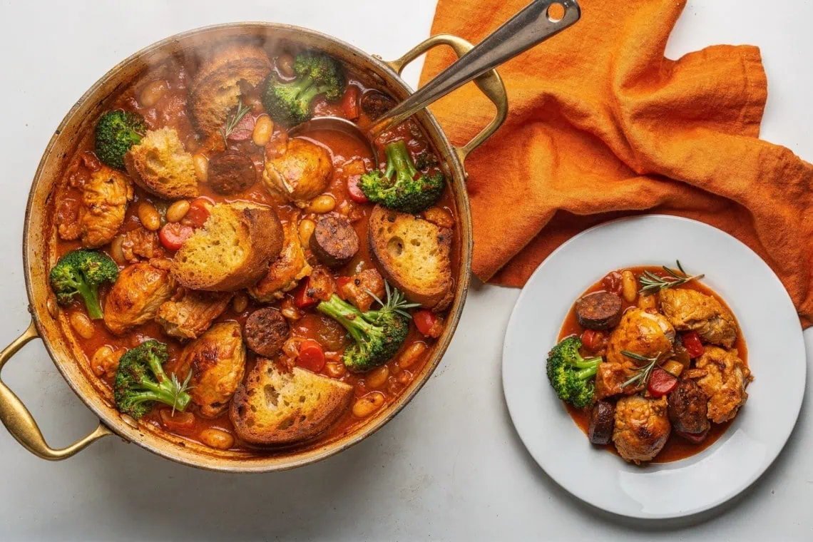 Quick Chicken, Sausage and Borlotti Bean Stew - Saffarazzi Hearty chicken stew with broccoli, beans, tomatoes and crusty bread in a copper pan, with an orange napkin nearby.