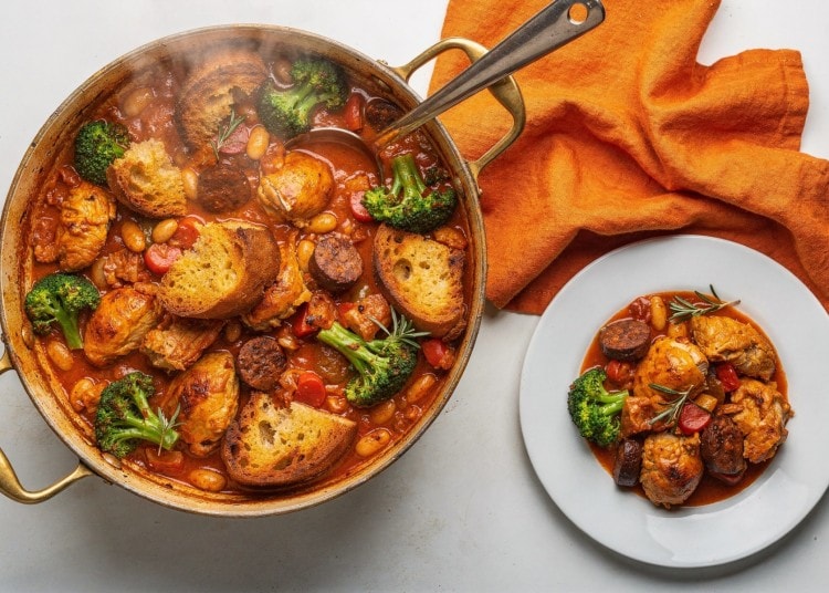 Hearty chicken stew with broccoli, beans, tomatoes and crusty bread in a copper pan, with an orange napkin nearby.