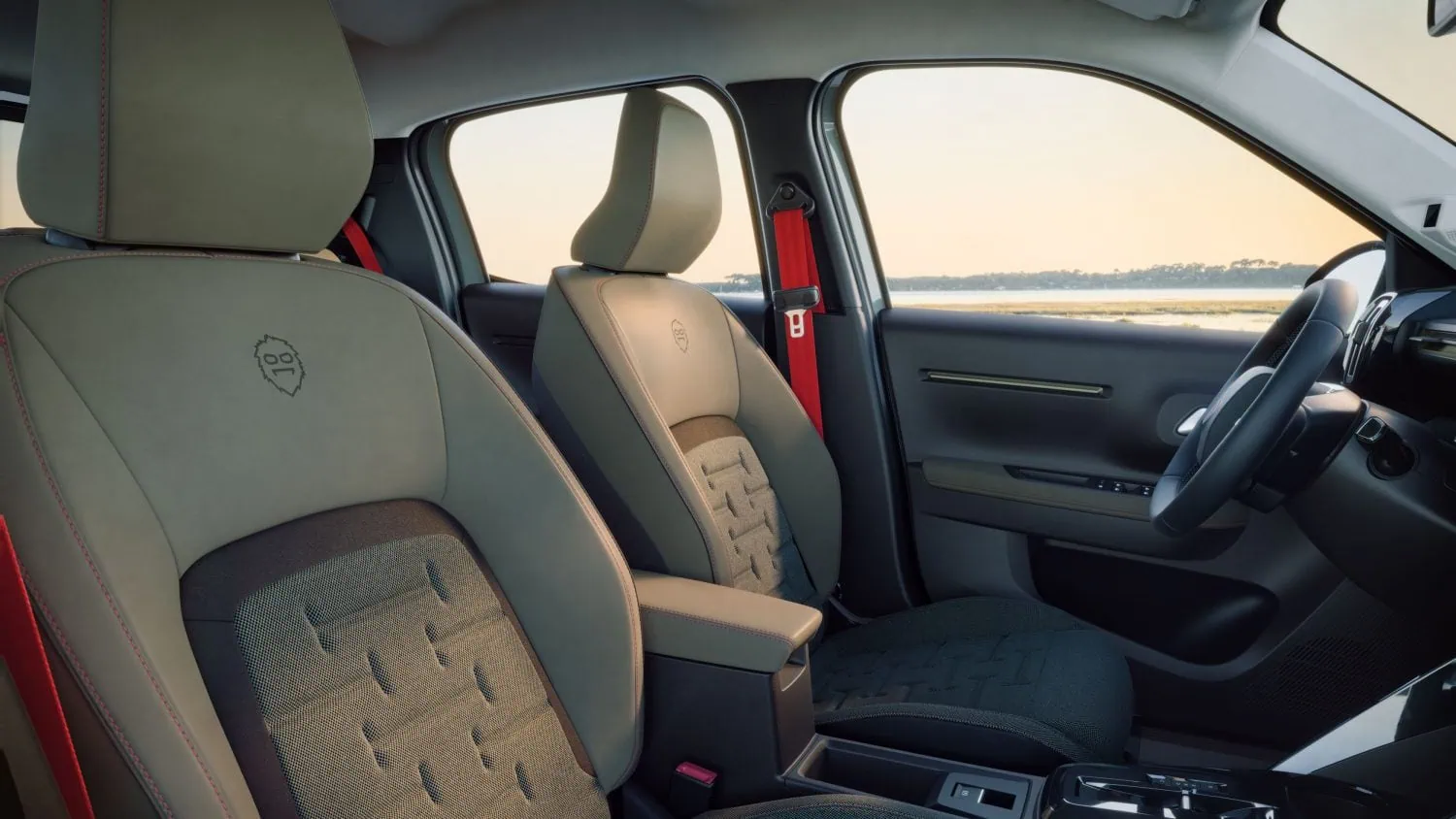 Citroën C3 OUTDOOR front seats with fabric upholstery - Saffarazzi Front interior of a car with beige seats and red seatbelts, overlooking a coastline through the windows.
