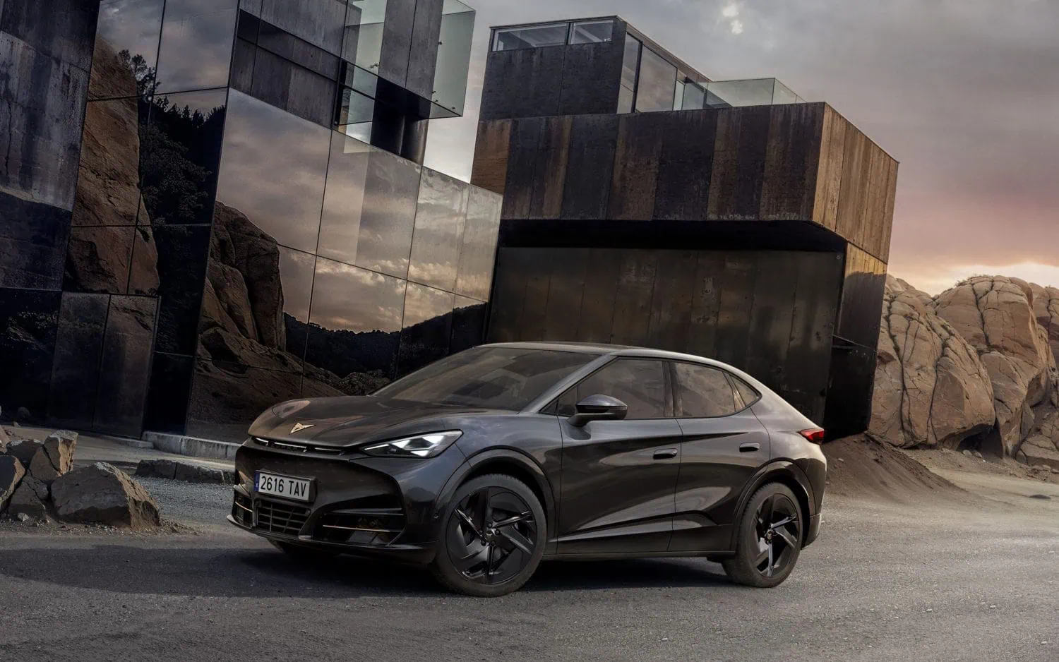 CUPRA Tavascan electric SUV beside modern building - Saffarazzi Gray luxury car with a sleek, low profile parked beside a modern glass-and-concrete building at dusk, with rocky terrain in the background.