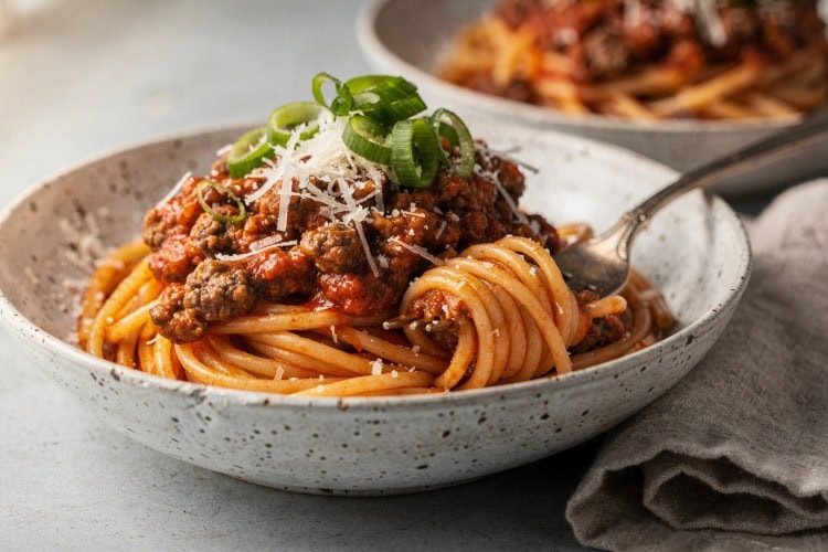 Easy spaghetti bolognese topped with grated Parmesan and sliced spring onions in a speckled white bowl
