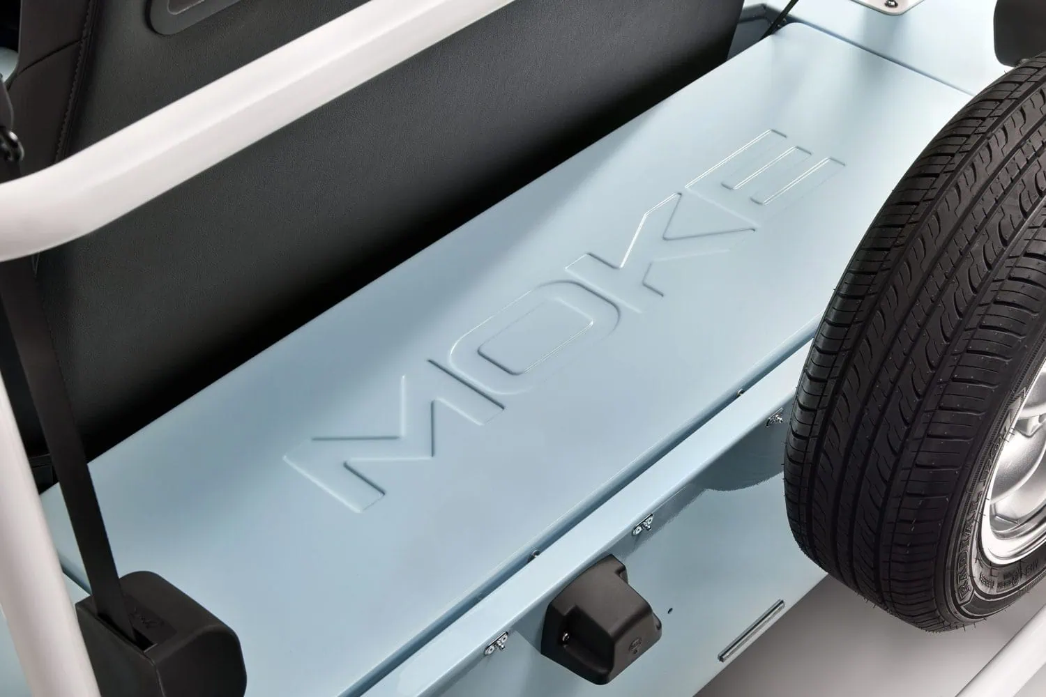 Electric MOKE Mk2 rear deck with MOKE lettering - Saffarazzi Open car trunk interior showing a light-blue cargo floor with molded panels and a spare tire along the right edge/styles metalwork nearby.