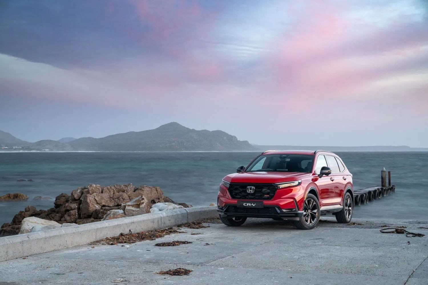 Honda CR-V hybrid SUV on slipway beside rocks - Saffarazzi Red Honda CR-V parked on a concrete jetty by the rocky shore, with calm sea, distant mountains, and a pastel sunset sky in the background.