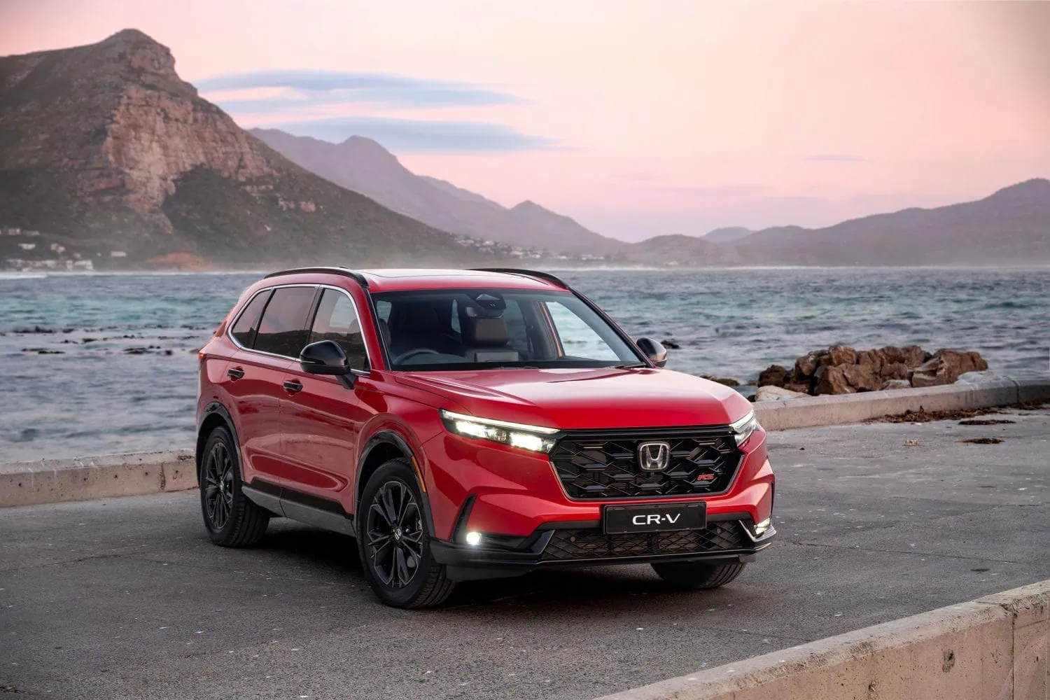 Honda CR-V hybrid SUV with mountains and sea behind - Saffarazzi Red Honda CR-V SUV parked along a coastal road with rocky breakwater and pastel sunset over mountains in the background.