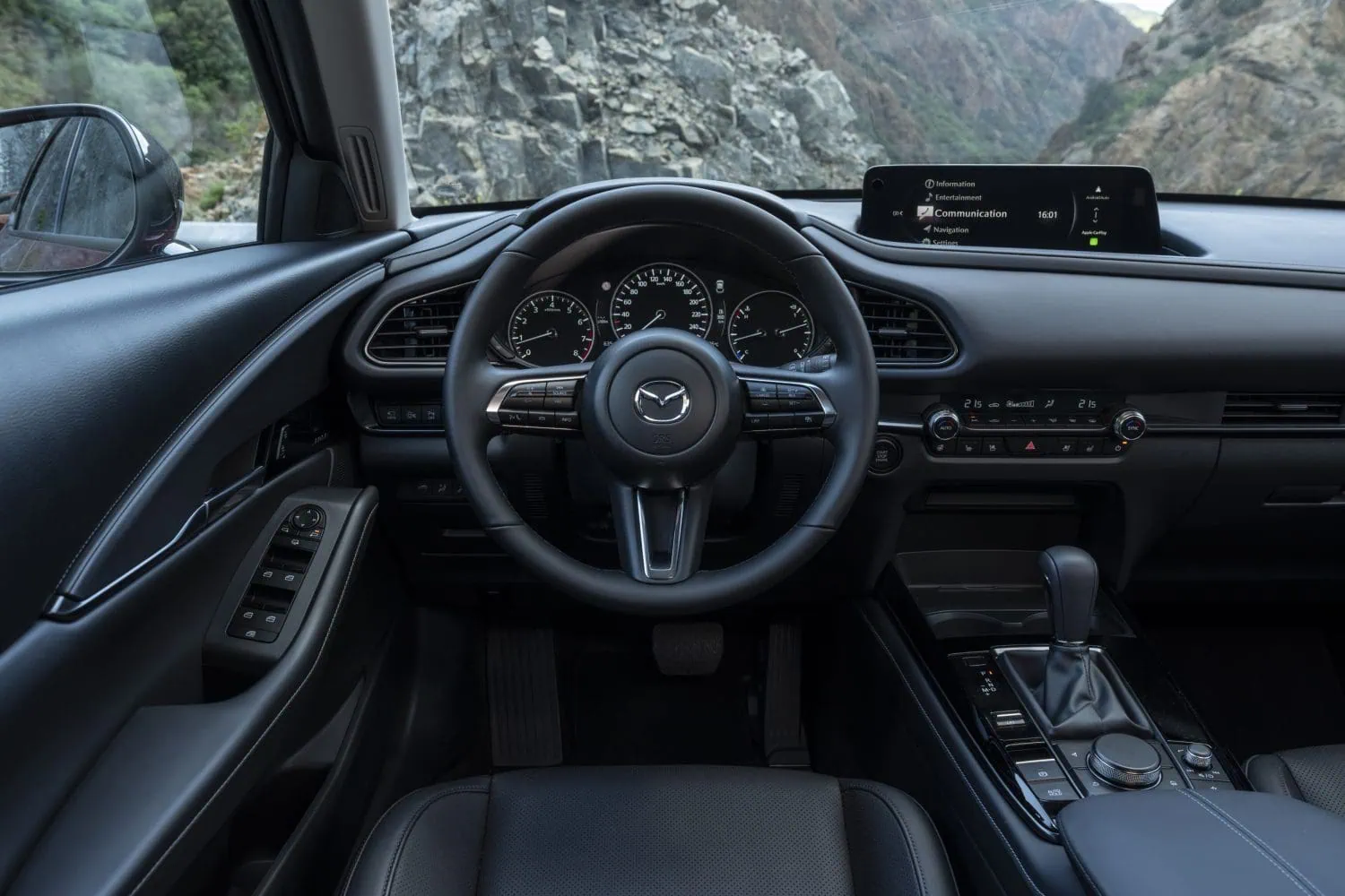 Mazda CX-30 dashboard and steering wheel - Saffarazzi Front interior of a Mazda SUV showing steering wheel, analog gauges, and a wide center touchscreen display with menus visible.