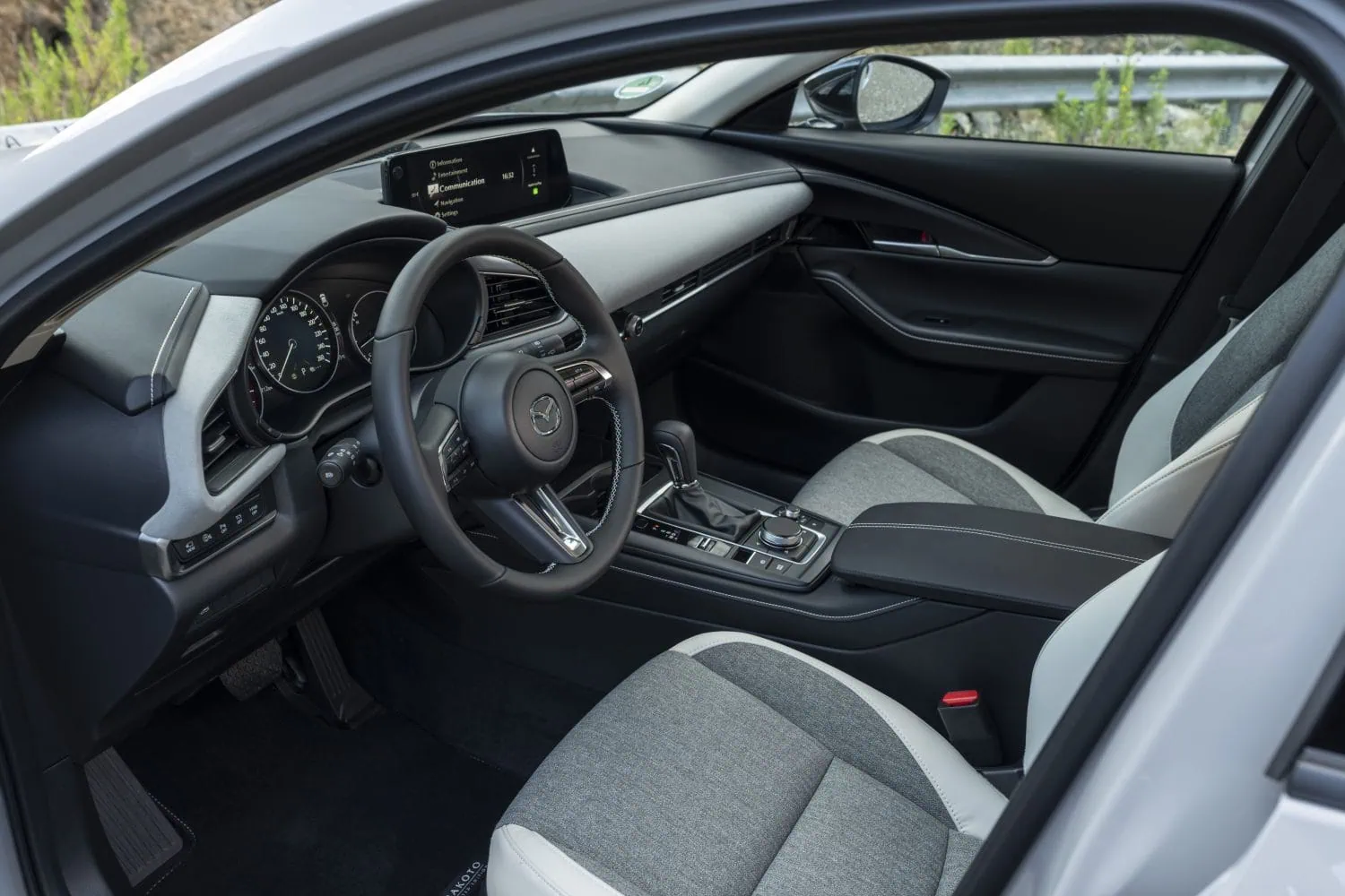 Mazda CX-30 interior with grey and white seats - Saffarazzi Interior view of a modern car showing a left-hand drive steering wheel, digital instrument cluster, center touchscreen, and black-gray upholstery with contrast stitching.