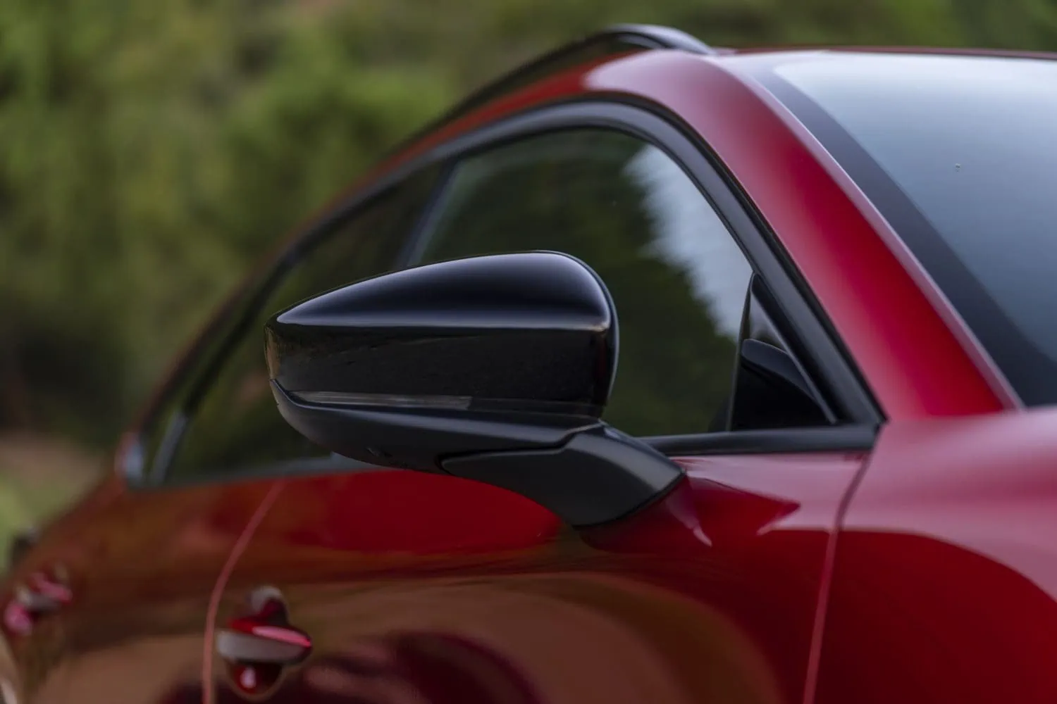 Red Mazda CX-30 black door mirror detail - Saffarazzi Close-up of a red car's glossy black side mirror beside a tinted window, with a blurred green background.