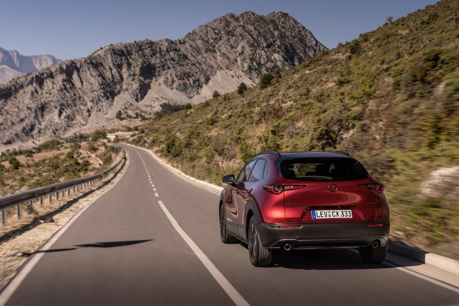 Red Mazda CX-30 rear view on a mountain road - Saffarazzi Red SUV on a winding mountain road with guardrails and steep rocky cliffs in the background, mountain scenery on a sunny day.