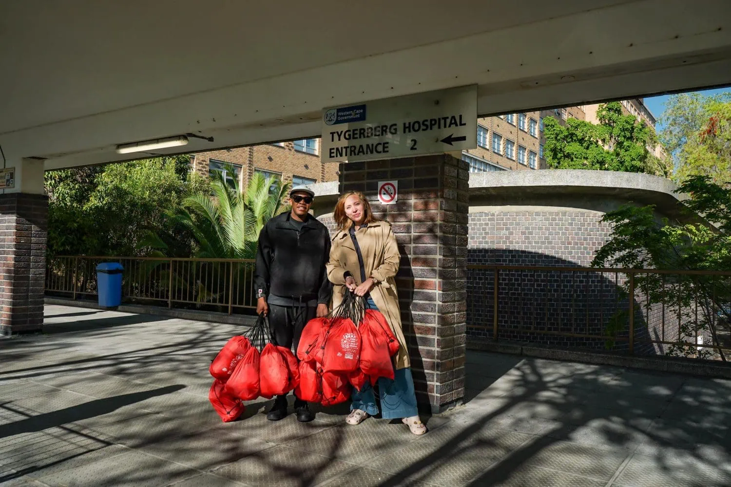 Seven7 Drive team with gift bags at Tygerberg Hospital - Saffarazzi Two people stand outside Tygerberg Hospital entrance with red bags in hand.