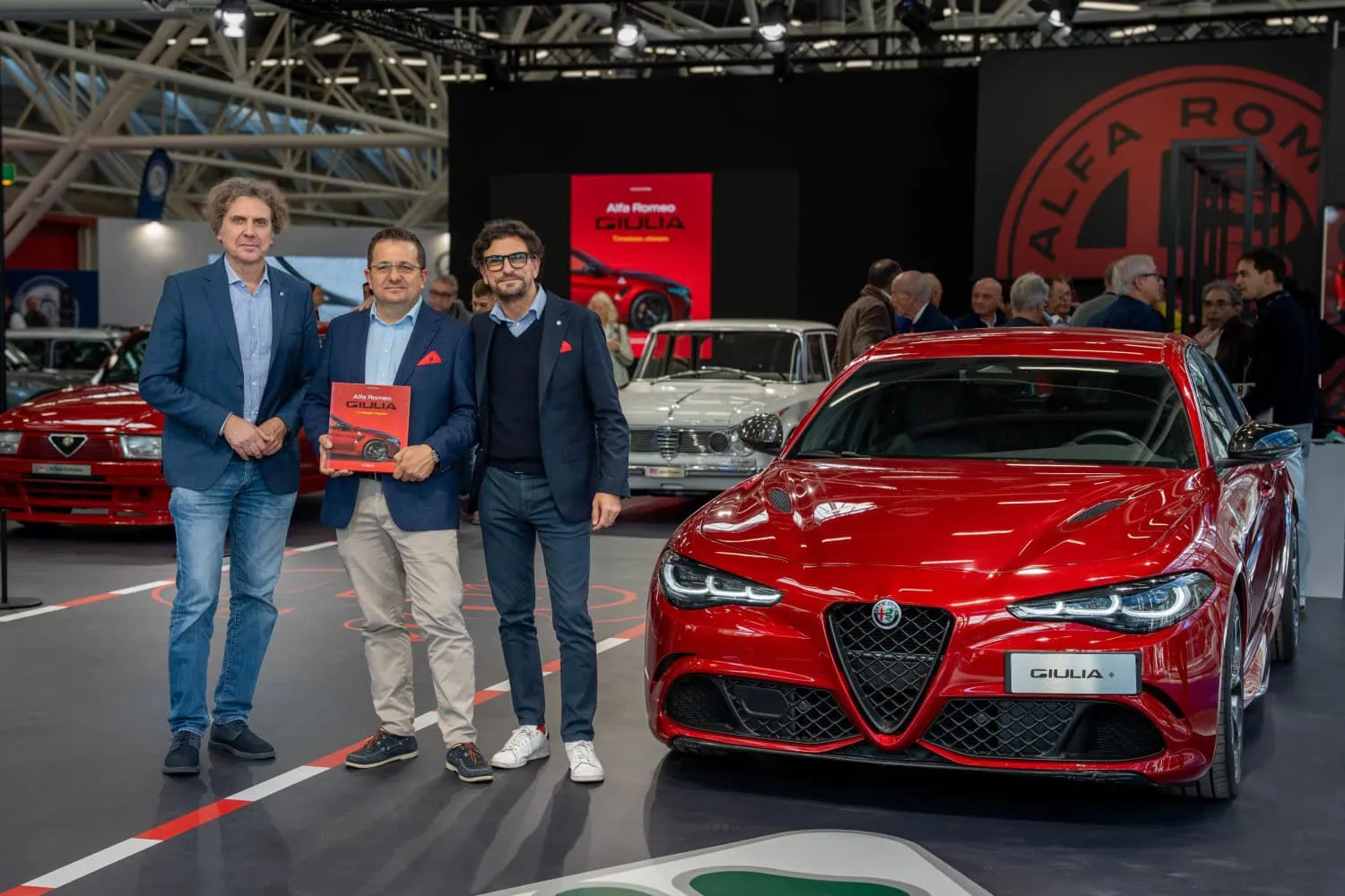 Three men with Giulia book - Saffarazzi Three men holding Giulia book beside Alfa Romeo Giulia.